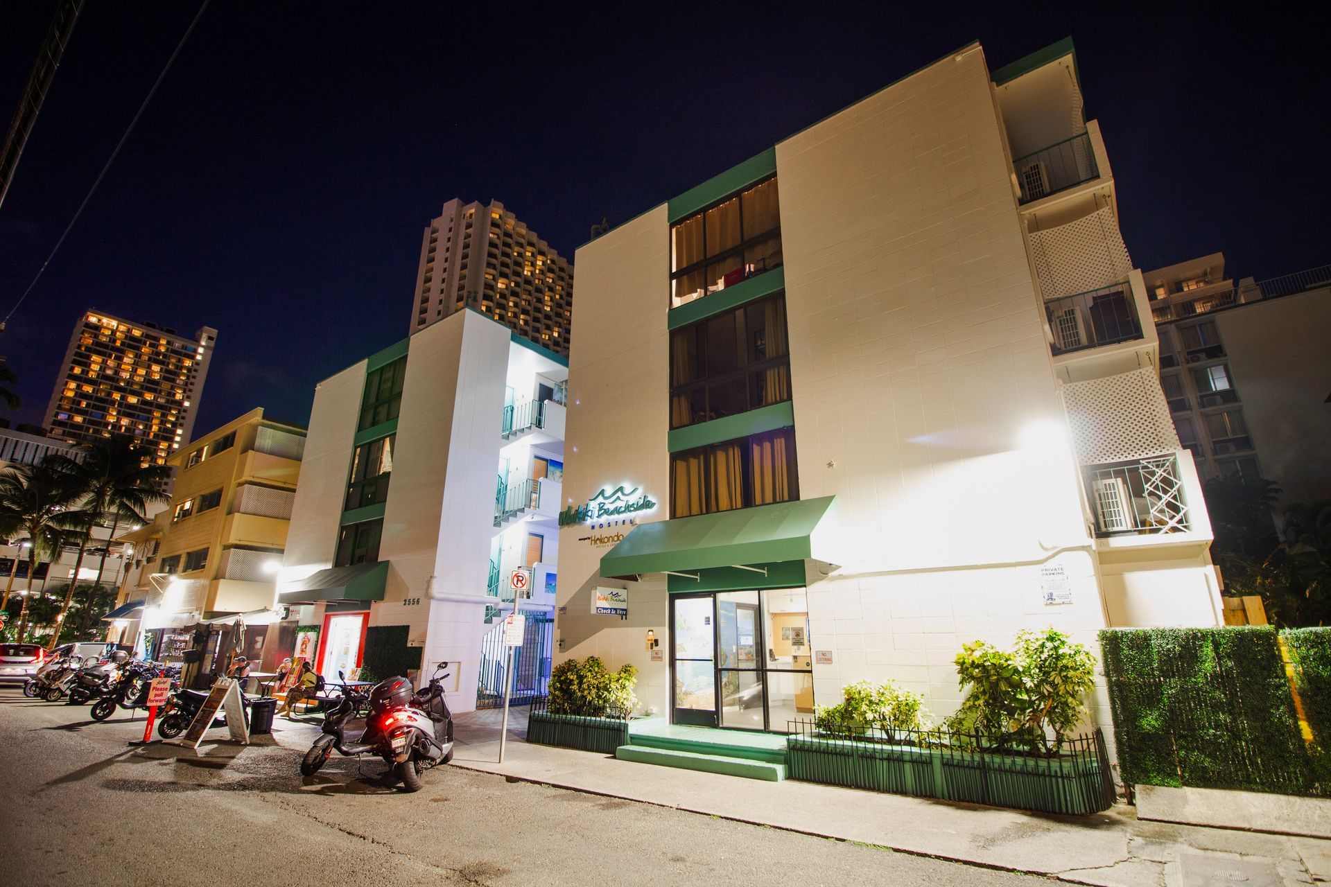 夜のホテル外観。白い建物にティール色のアクセント。通りにはバイクが停められている。