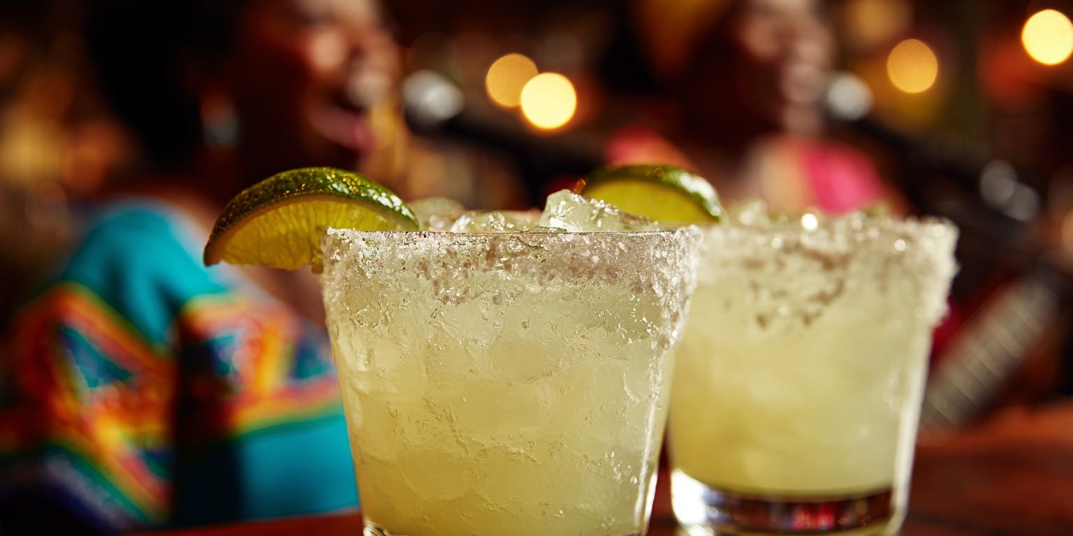 Two margaritas are sitting on a table with a woman in the background.