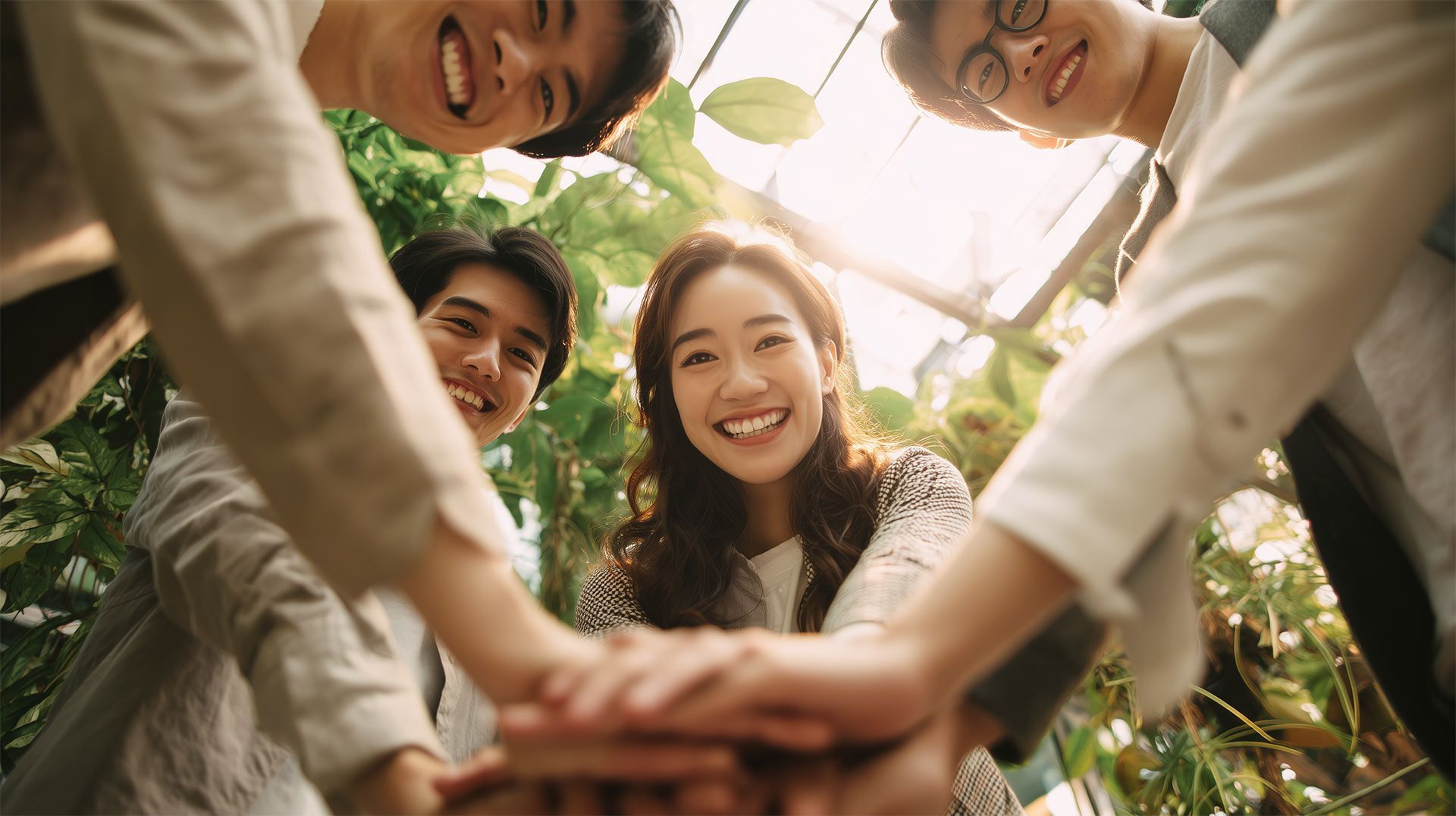 Four smiling young adults, hands stacked together, teamwork, sunny setting.